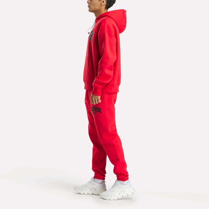 A person in profile wears a bright red Reebok Men's Hoodie with matching sweatpants featuring black text, paired with white sneakers, all set against a plain white background.