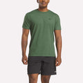 A man wearing a green Reebok Men's Identity Small Logo T-Shirt and black Reebok shorts stands facing forward against a plain, light background.