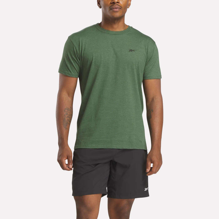 A man wearing a green Reebok Men's Identity Small Logo T-Shirt and black Reebok shorts stands facing forward against a plain, light background.