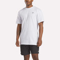 A man wears a relaxed fit white Reebok Men's Identity Small Logo T-Shirt and black Reebok shorts, standing against a white background.