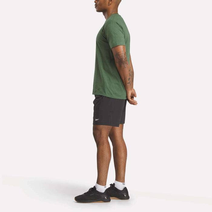 A person stands in profile facing left, wearing a green Reebok Men's Identity Small Logo T-Shirt, black athletic shorts, black sneakers, and white socks with hands clasped behind their back against a plain white background.