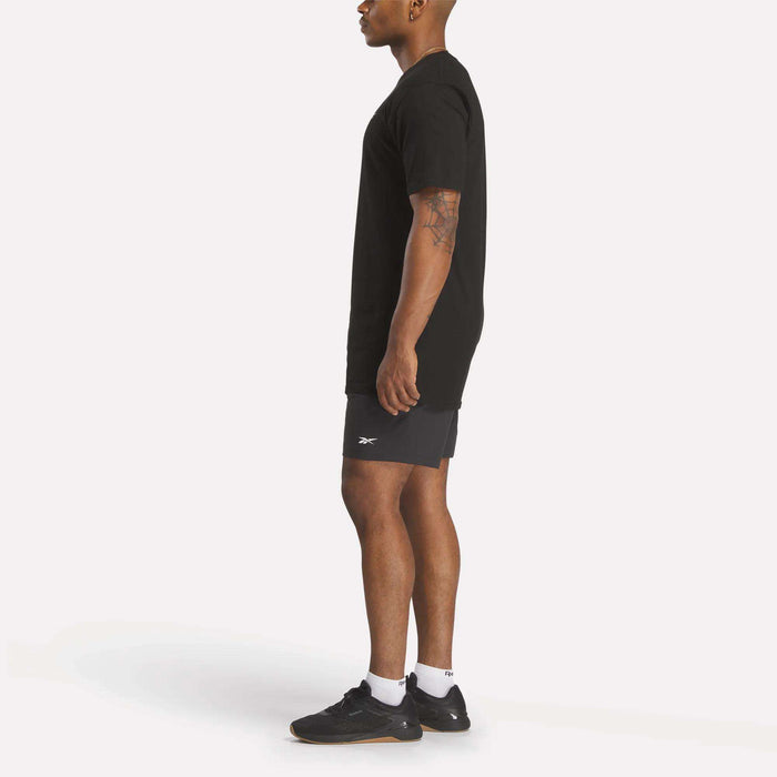 A person in profile wears a relaxed fit black Reebok Men's Identity Small Logo T-Shirt, dark athletic shorts, white socks, and black Reebok sneakers against a plain white background.