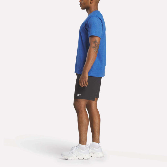 A man stands in profile wearing a relaxed fit blue Reebok Men's Identity Small Logo T-Shirt, black athletic shorts, and white Reebok sneakers. He has an upper left arm tattoo and stands against a plain white background.
