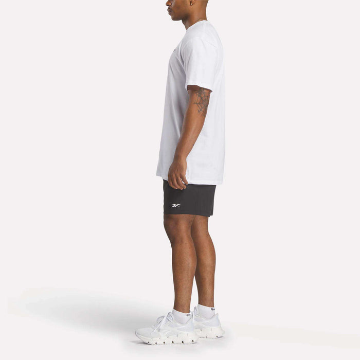 A man stands in profile against a white background wearing a relaxed fit Reebok Men's Identity Small Logo T-Shirt, black Reebok shorts, and white athletic shoes.