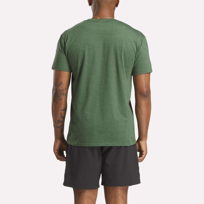 A person stands with their back to the camera, wearing a green Reebok Men's Identity Small Logo T-Shirt in a relaxed fit and black shorts. Their arms are tattooed, and they are posed against a plain light background.