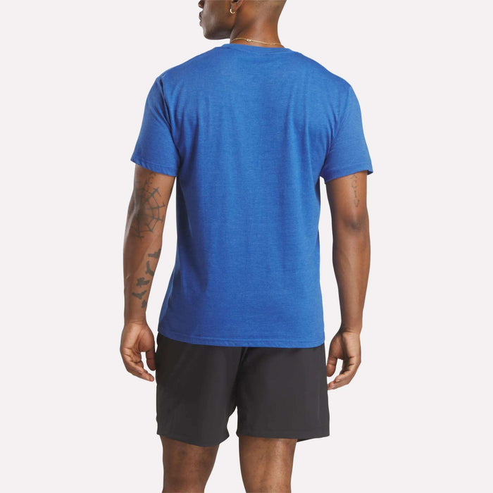 A person faces away, wearing a blue Reebok Men's Identity Small Logo T-Shirt with a relaxed fit and black shorts. Their arms are by their sides, showing tattoos on the left arm. The background is plain and light-colored.
