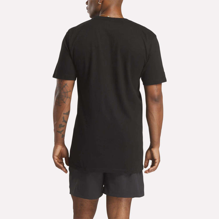 A person with tattoos on their left arm stands facing away from the camera in a relaxed fit Reebok Men's Identity Small Logo T-Shirt and black shorts. The background is plain and light-colored, showcasing the back of the outfit.