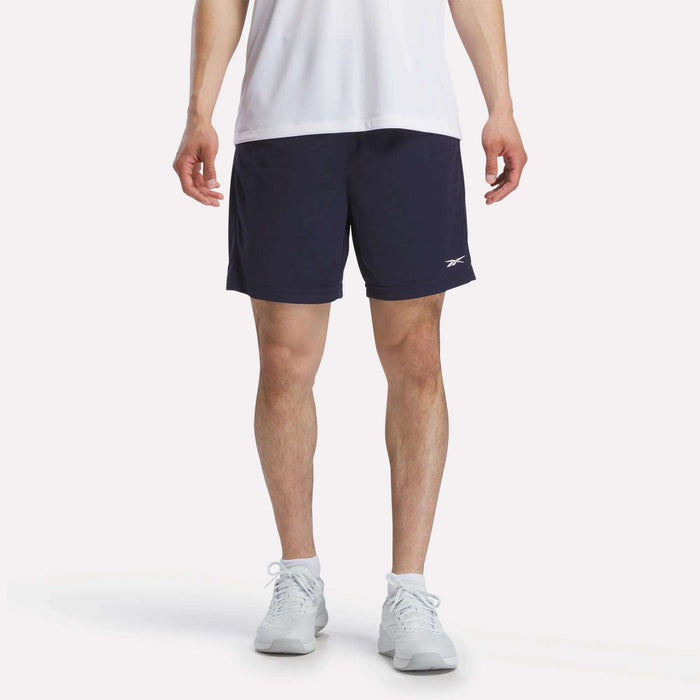 A person wearing a white T-shirt, white socks, and white sneakers is standing against a plain white background, paired with Reebok Men's Inclination Shorts in navy blue featuring a small logo.