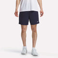 A person wearing a white T-shirt, white socks, and white sneakers is standing against a plain white background, paired with Reebok Men's Inclination Shorts in navy blue featuring a small logo.