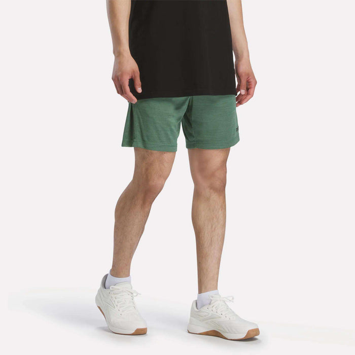 A person stands against a plain white background wearing a black t-shirt, white socks, white sneakers, and green Reebok Men's Inclination Shorts.