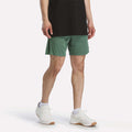A person stands against a plain white background wearing a black t-shirt, white socks, white sneakers, and green Reebok Men's Inclination Shorts.