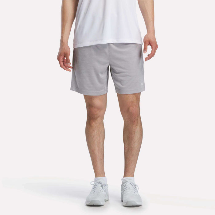A person wears a white T-shirt, Reebok Men's Inclination Shorts in light gray, and white sneakers while facing forward against a plain white background. The image is cropped at the shoulders.