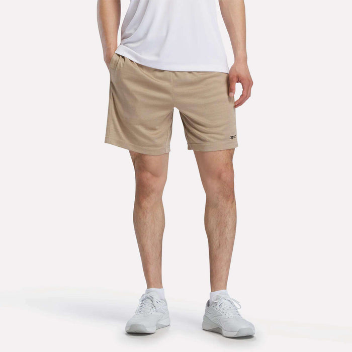 A person stands against a plain white background, showing only their lower torso and legs. They are wearing white sneakers, a white T-shirt, and Reebok Men's Inclination Shorts.