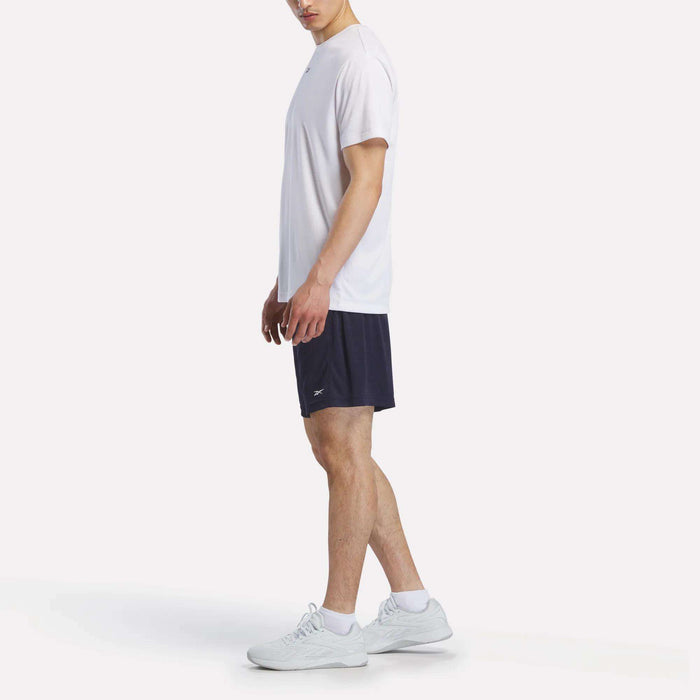 A man stands sideways wearing a plain white t-shirt, Reebok Men's Inclination Shorts, and white sneakers against a plain light background.