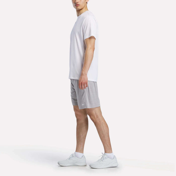 A person stands sideways against a plain, light background wearing a white t-shirt, white sneakers, and Reebok Men's Inclination Shorts.