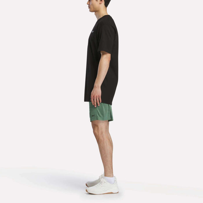 A person stands in profile against a plain white background, wearing a black t-shirt, Reebok Men's Inclination Shorts in green, and white athletic shoes.