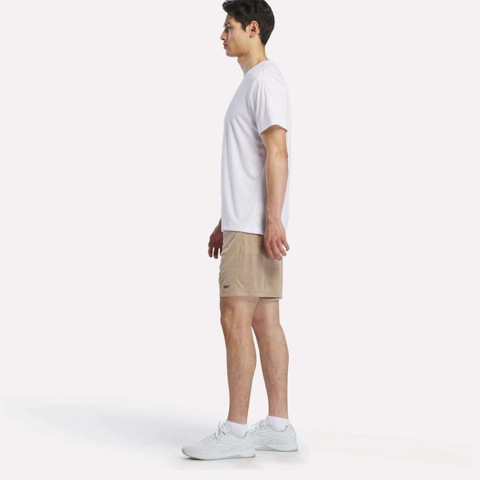 A man stands in profile against a white background, wearing a white T-shirt, Reebok Men's Inclination Shorts, and white sneakers.