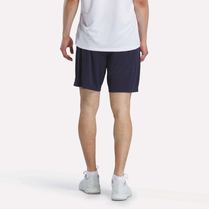 A person stands with their back to the camera, wearing a white shirt, Reebok Men's Inclination Shorts in navy blue, and white athletic shoes against a plain white background.