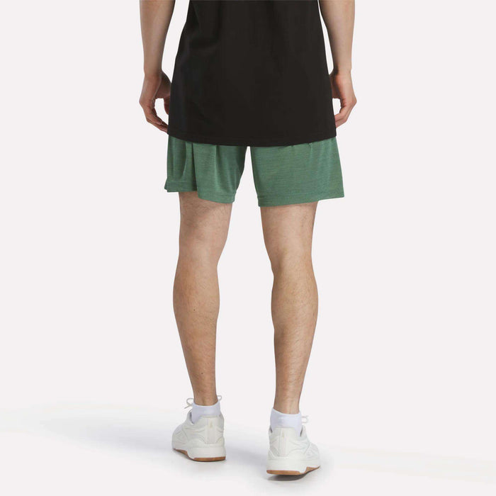 A person stands with their back to the camera, wearing a black shirt, white sneakers, and green Reebok Men's Inclination Shorts against a plain white background.