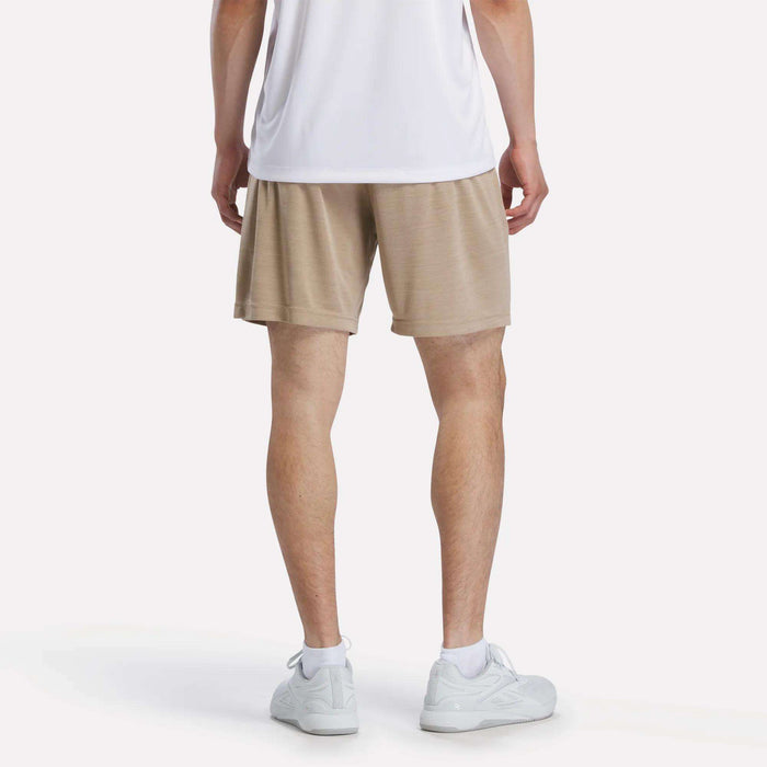 A person stands with their back to the camera in a white shirt, white sneakers, and tan Reebok Men's Inclination Shorts against a plain white background.