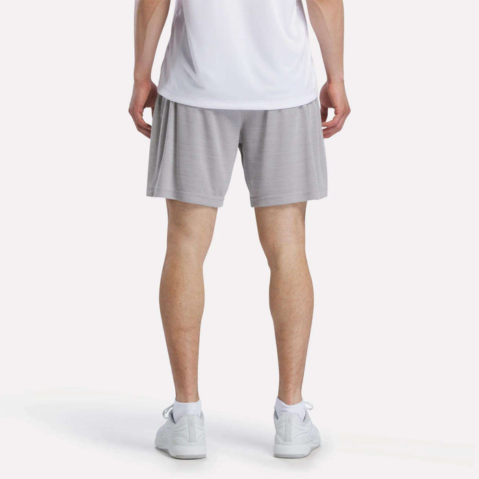 A person is shown from the waist down against a plain light background, wearing a white shirt, Reebok Men's Inclination Shorts in light gray, and white sneakers.