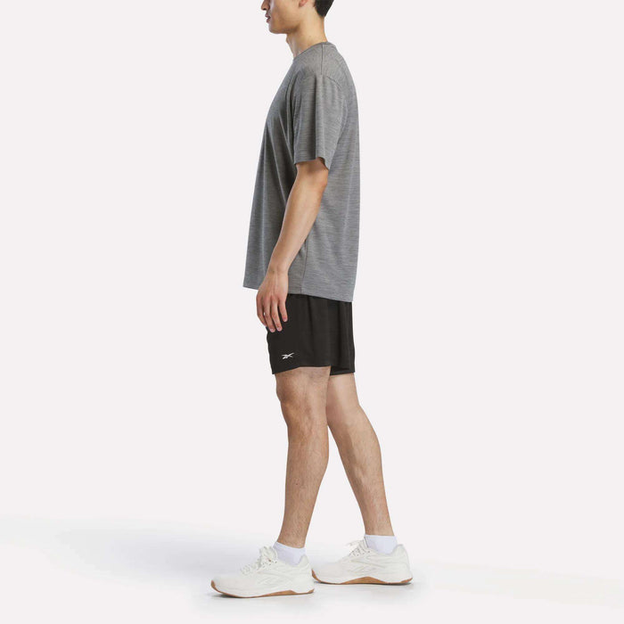A man stands in profile wearing a loose grey t-shirt, Reebok Men's Inclination Shorts in black, and white athletic shoes against a plain white background.