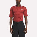 A man stands facing forward against a white background wearing a brick red/chalk Men's Jacquard Polo Shirt from Reebok, featuring a white logo on the chest, paired with a black belt and black shorts.
