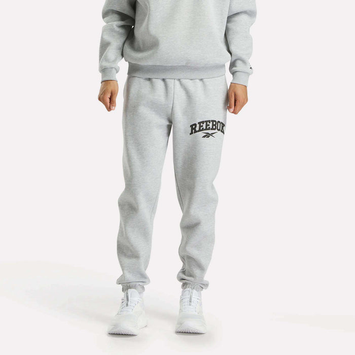 A person models Reebok Men's Joggers in light gray with a black logo on the left thigh, styled with a matching sweatshirt and white sneakers against a plain white background.