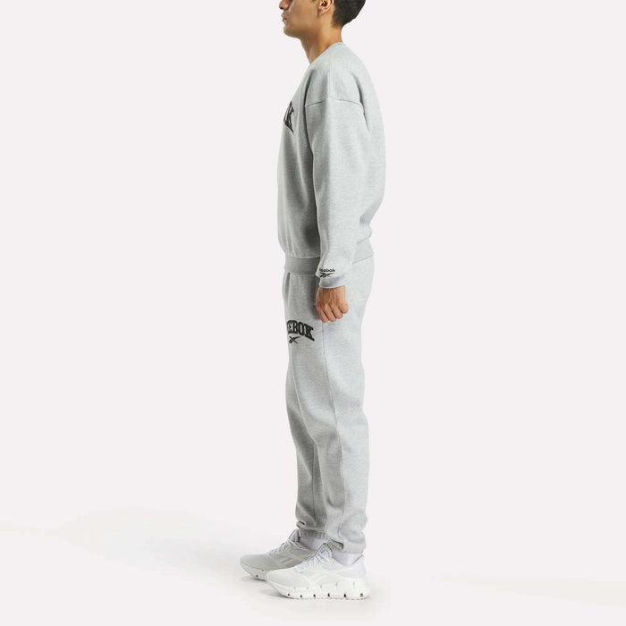 A person stands in profile wearing Reebok Men's Joggers with a matching light gray sweatshirt and white sneakers, set against a plain white background.
