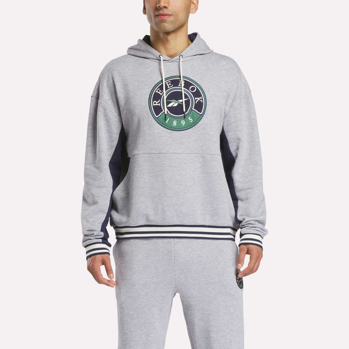 A man stands against a plain white background wearing the Reebok Men's Lawn Club French Terry Hoodie in light gray with a green and navy logo, paired with matching sweatpants. This cotton blend hoodie offers comfort and style.