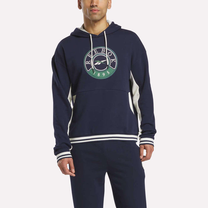 A person wears the Reebok Men's Lawn Club French Terry Hoodie in navy blue, featuring white accents and a green, white, and navy Reebok logo on the chest, paired with matching navy pants against a plain light background.