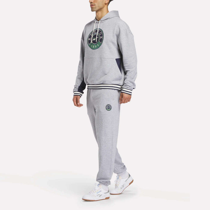 A person wearing the Reebok Men's Lawn Club French Terry Hoodie in light gray and matching sweatpants, both featuring the round Reebok logo, along with white athletic shoes, stands against a plain white background.