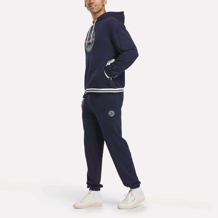 A person smiles in a Reebok Men's Lawn Club French Terry Hoodie—navy blue with white trim and green circular logo—matched with coordinated sweatpants and white sneakers, posing against a plain light background.