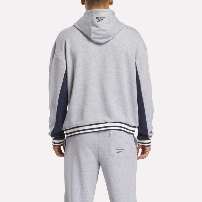 A person wearing a light gray Reebok Men's Lawn Club French Terry Hoodie, seen from behind. This hoodie features dark blue sleeve accents and striped cuffs and waistband.