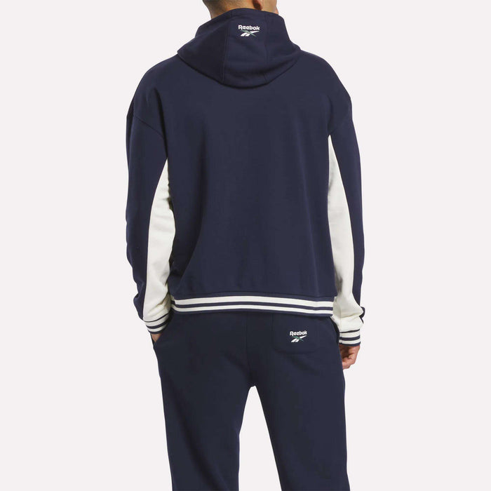 A person faces away wearing the Reebok Men's Lawn Club French Terry Hoodie tracksuit in navy blue with white sleeve stripes and trim. The cotton-blend hoodie displays the Reebok logo on the hood's back and pants pocket.