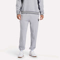 A person in Reebok Men's Lawn Club French Terry Jogger Pants and a matching gray sweatshirt with black and white striped cuffs and hem, paired with white sneakers, stands against a plain white background.