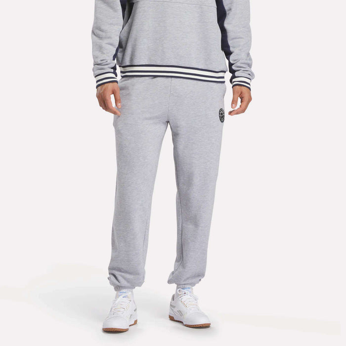 A person in Reebok Men's Lawn Club French Terry Jogger Pants and a matching gray sweatshirt with black and white striped cuffs and hem, paired with white sneakers, stands against a plain white background.