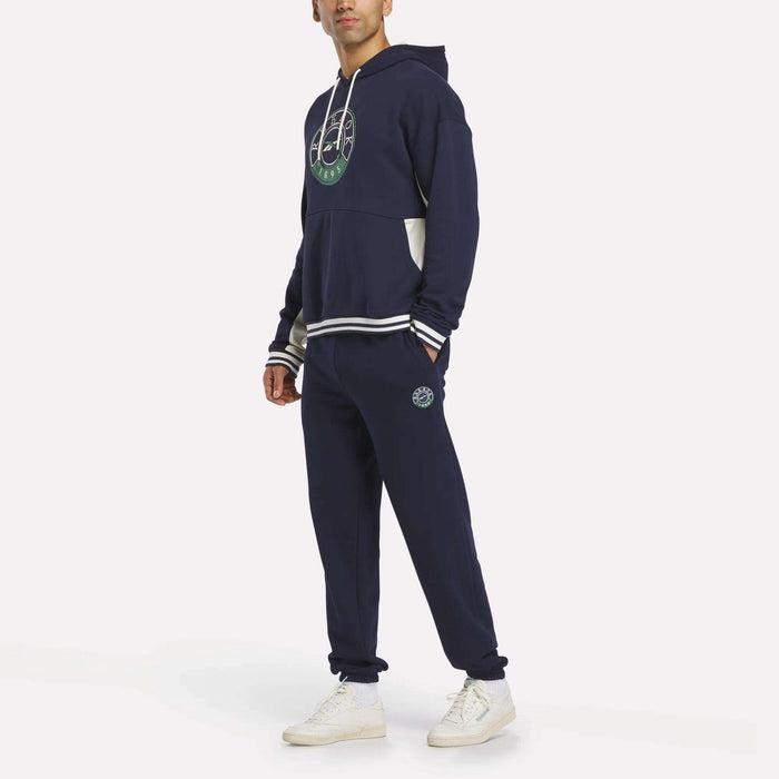 A person stands against a plain white background wearing a navy blue hoodie with a green circular graphic, Reebok Men's Lawn Club French Terry Jogger Pants, and white sneakers.