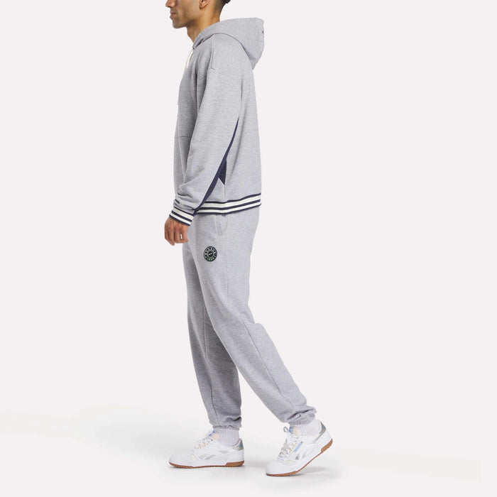A man in a grey Reebok hooded tracksuit and white sneakers stands sideways against a white background, modeling the Men's Lawn Club French Terry Jogger Pants for a classic sporty look.