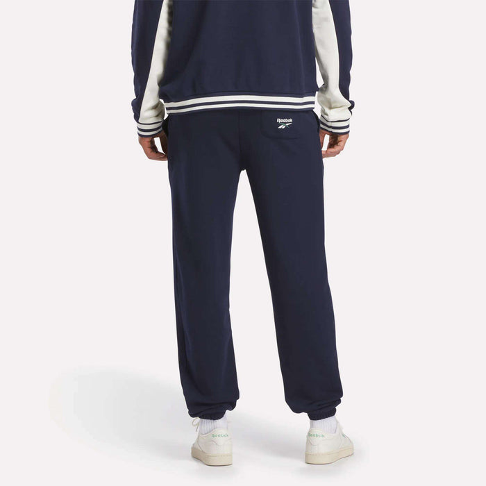 A person stands facing away, wearing Reebok Men's Lawn Club French Terry Jogger Pants in navy with a white logo on the back pocket, white sneakers, and a matching navy jacket—classic sporty style crafted from a comfortable cotton-blend.