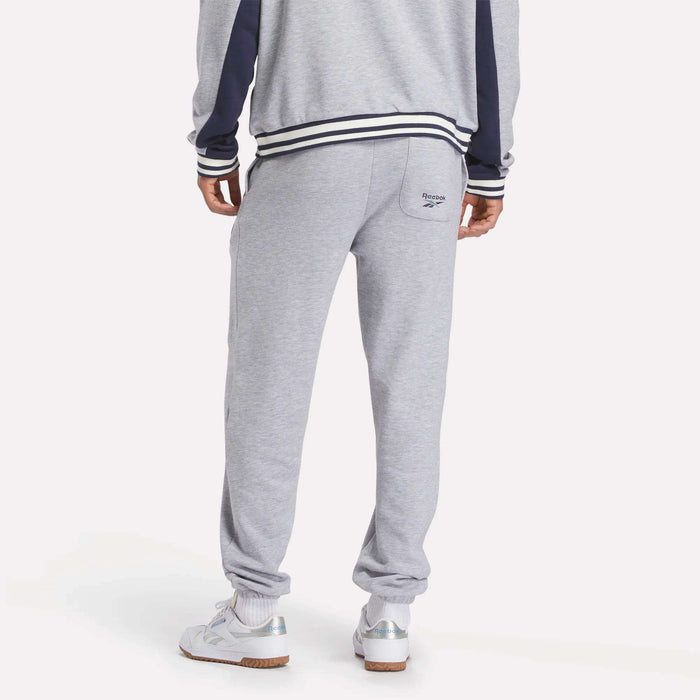 A person is shown from the back wearing Reebok Men's Lawn Club French Terry Jogger Pants, a grey tracksuit with navy sleeve stripes and a small logo on the right pocket, paired with white sneakers.