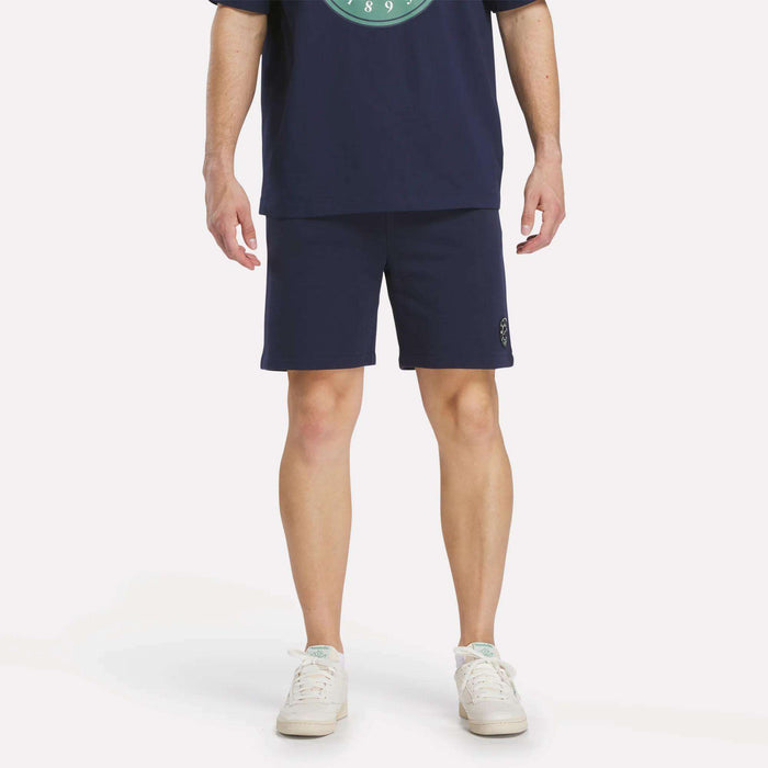 A person stands with arms relaxed by their sides, wearing a navy blue T-shirt, Reebok Men's Lawn Club Shorts, and white sneakers. The image is cropped at the shoulders, so their face is not visible.