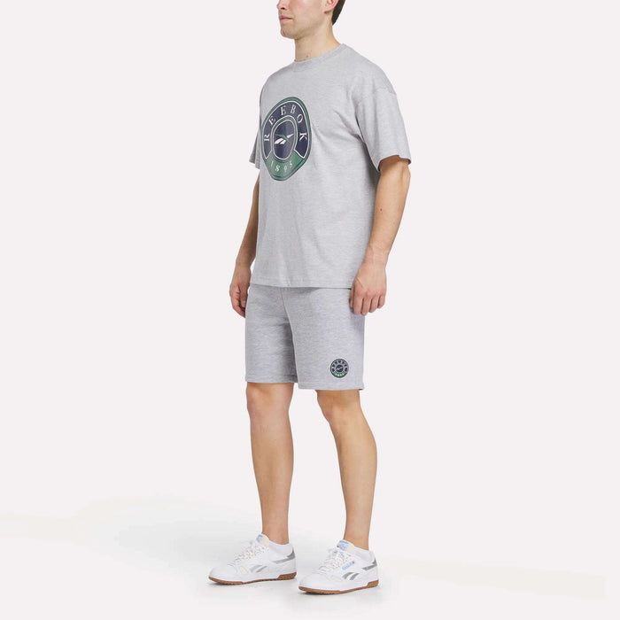 A person wearing a light grey T-shirt with a circular logo, Reebok Men's Lawn Club Shorts, and white sneakers stands against a plain white background.