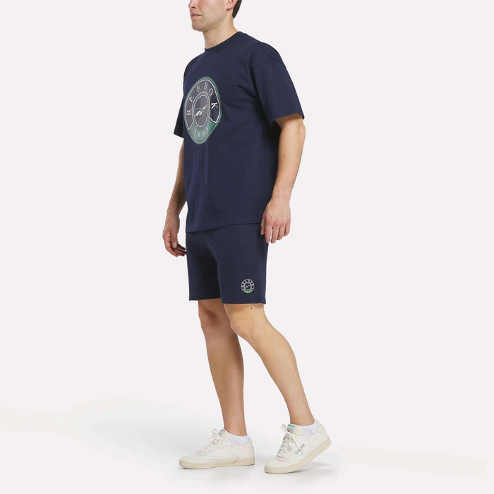 A person in a navy blue t-shirt with circular graphics, Reebok Men's Lawn Club Shorts featuring similar designs, and white sneakers stands against a plain white background.