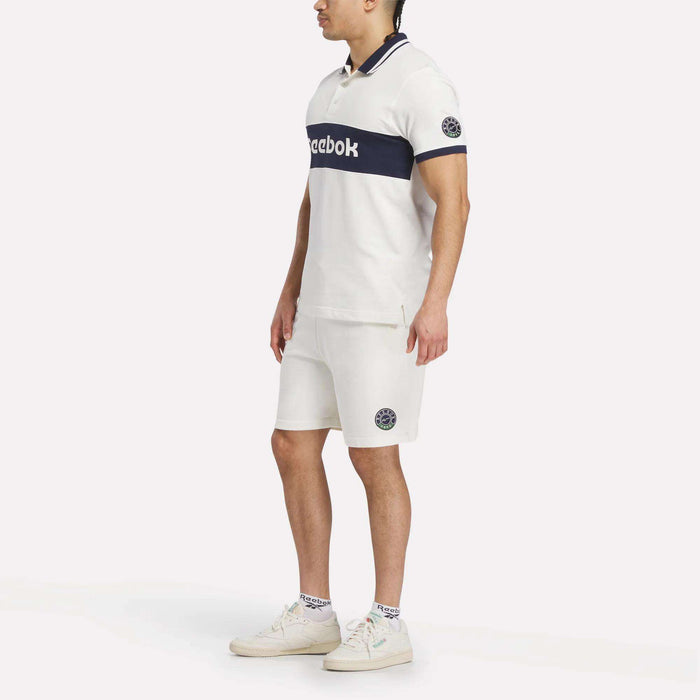 A man stands on a plain light background wearing Reebok’s Men's Lawn Club Shorts with navy blue accents, a matching white Reebok polo shirt, white Reebok sneakers, and white socks—all featuring the iconic Reebok logo.