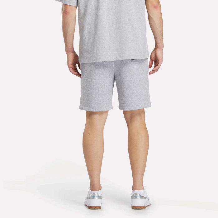 A person stands with their back to the camera, wearing a light gray T-shirt, white sneakers, and Reebok Men's Lawn Club Shorts. The plain, light background highlights the shorts' casual style.
