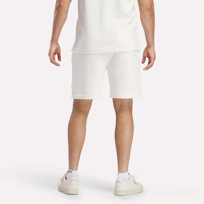 A person in a white t-shirt, Reebok Men's Lawn Club Shorts, and white sneakers stands with their back to the camera against a plain white background.