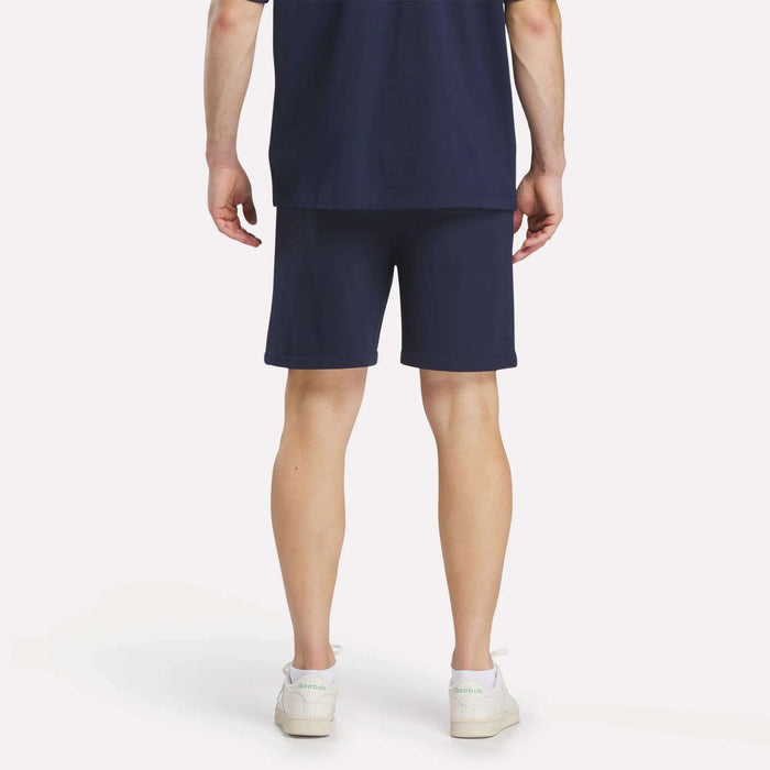 A person faces away, wearing a navy blue T-shirt, Reebok Men's Lawn Club Shorts, and white sneakers against a plain white background.