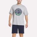 A man in a light gray Reebok Men's Lawn Club T-Shirt with a large circular logo and navy blue shorts stands facing forward against a plain white background.
