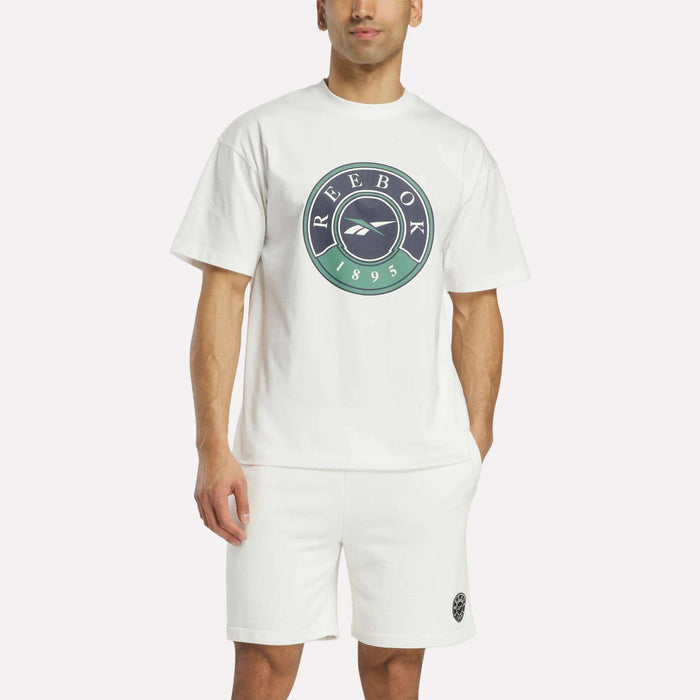 A man wearing Reebok's Men's Lawn Club T-Shirt, a soft cotton tee with a large circular logo, and matching white shorts with a small emblem stands against a plain light background.